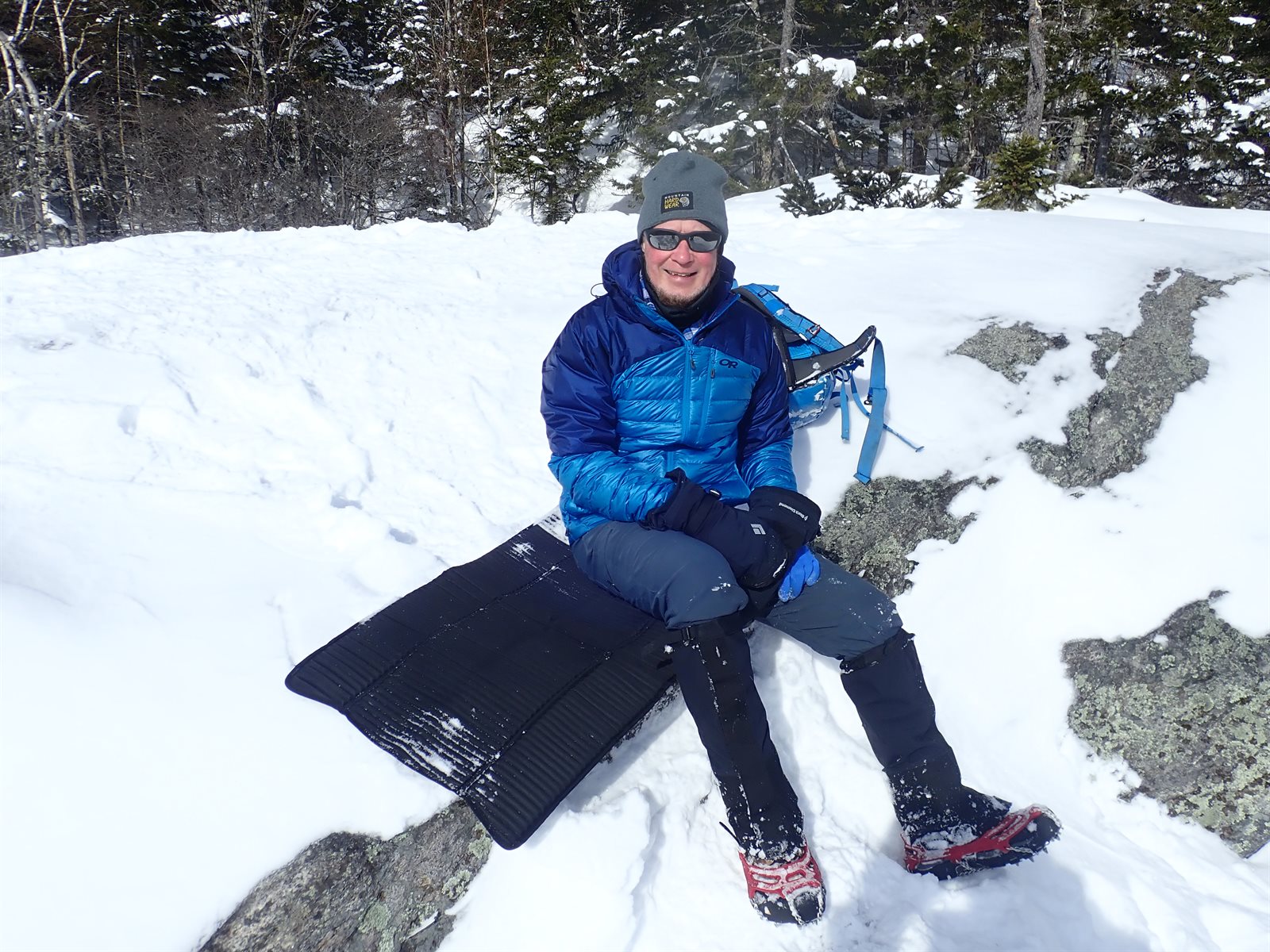 A hiker in a warm puffy jacket resting on an insulated sit pad on a snowy winter summit